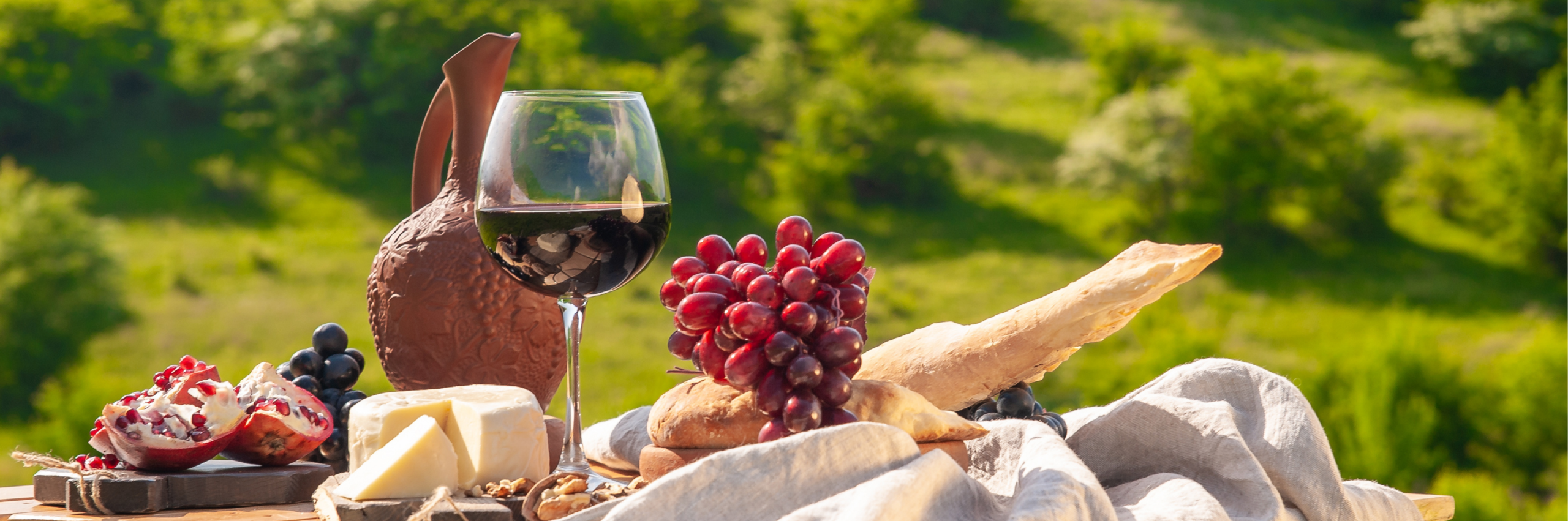 Tavola imbandita all’aperto con vino rosso, uva, formaggi, pane e frutta, con paesaggio verde sullo sfondo